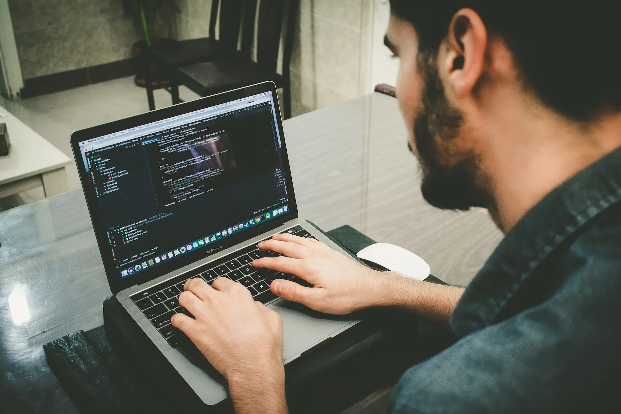 Person typing code on a MacBook Pro laptop at a desk with a white Apple Magic Mouse beside it.